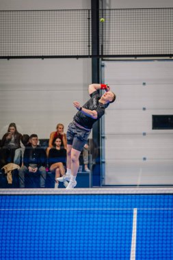 Riga, Letonya - 24 Ocak 2026: Padel turnuvası. Gri spor giyimli bir erkek sporcu, kapalı bir padel sahasında seyircilerin izlediği bir topa servis yapıyor.