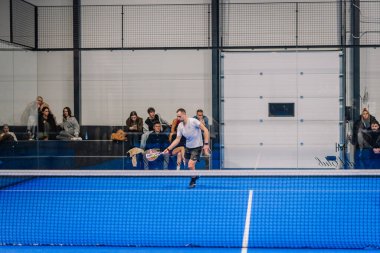 Riga, Letonya - 24 Ocak 2026: Padel turnuvası. Mavi kortta padel oynayan erkek sporcu, bir spor tesisinin arka planında seyirciler oturuyor.