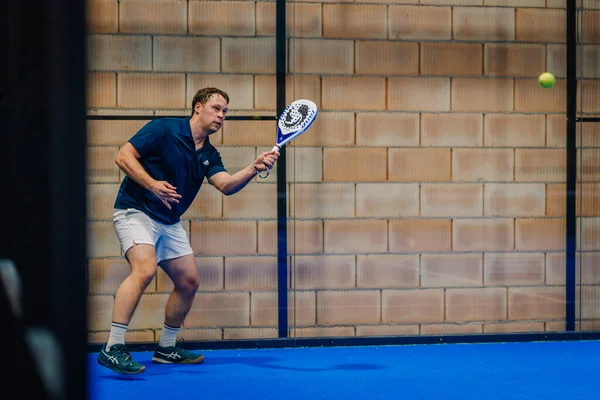 Riga, Letonya - 24 Ocak 2026: Padel turnuvası. Mavi tişörtlü ve beyaz şortlu bir erkek sporcu kapalı sahada topa vuracak pozisyonda.