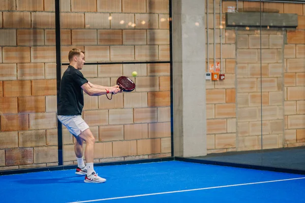 Riga, Letonya - 24 Ocak 2026: Padel turnuvası. Siyah tişörtlü ve beyaz şortlu erkek sporcu, kapalı alanda mavi bir kortta raketle padel topuna vuruyor.