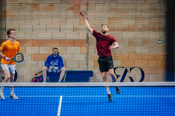Riga, Letonya - 24 Ocak 2026: Padel turnuvası. Kırmızı tişörtlü erkek atlet, turuncu tişörtlü takım arkadaşı iç sahada oynamaya hazırlanırken padel topu servis ediyor.