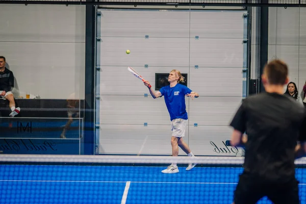 Riga, Letonya - 24 Ocak 2026: Padel turnuvası. Mavi tişörtlü ve beyaz şortlu erkek padel oyuncusu, cam duvarlı bir kortta topa vurmaya hazırlanıyor.