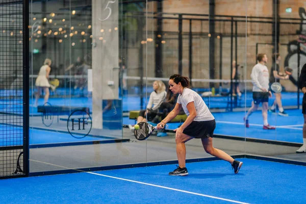 Riga, Letonya - 24 Ocak 2026: Padel turnuvası. Beyaz tişörtlü ve siyah şortlu bir bayan atlet, padel sahasında cam duvarlarla topa vurmaya hazırlanıyor.