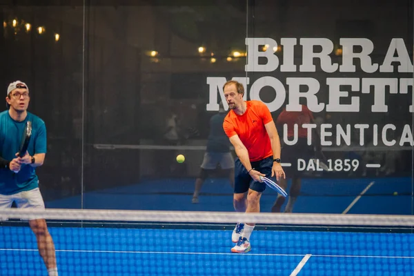 Riga, Letonya - 24 Ocak 2026: Padel turnuvası. İki erkek Padel oyuncusu mavi kortta bir maç yapıyor, biri turuncu tişört giyiyor, diğeri mavi.
