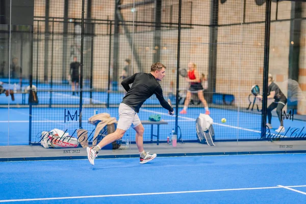 Riga, Letonya - 24 Ocak 2026: Padel turnuvası. Siyah elbiseli erkek padel oyuncu, arka planda cam duvarlar ve diğer oyuncularla mavi bir sahada topa vuruyor.