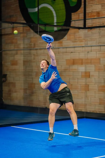 Riga, Letonya - 24 Ocak 2026: Padel turnuvası. Mavi tişörtlü erkek padel oyuncusu yeşil logolu mavi bir sahada topa servis yapmaya hazırlanıyor.