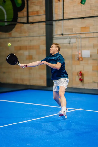 Riga, Letonya - 24 Ocak 2026: Padel turnuvası. Mavi tişörtlü ve beyaz şortlu erkek padel oyuncusu mavi kortta dokulu bir duvarla topa vuruyor
