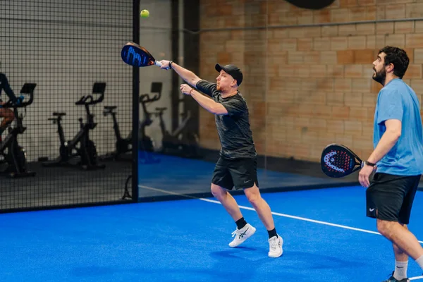 Riga, Letonya - 24 Ocak 2026: Padel turnuvası. Biri siyah tişörtlü ve şapkalı iki adam içeride Padel oynuyor. Topa vurmaya hazırlanıyor, diğeri mavi gömlekli.