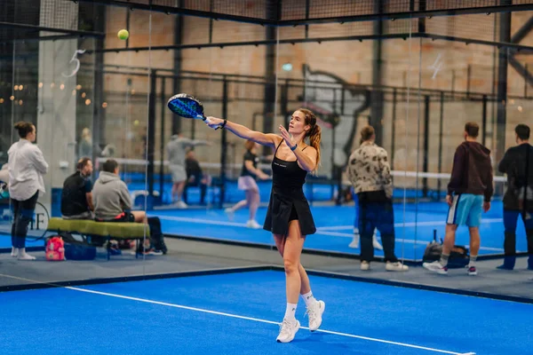 Riga, Letonya - 24 Ocak 2026: Padel turnuvası. Siyah elbiseli kadın padel oyuncu, kapalı bir sahada seyircilerle birlikte mavi raket sallıyor.