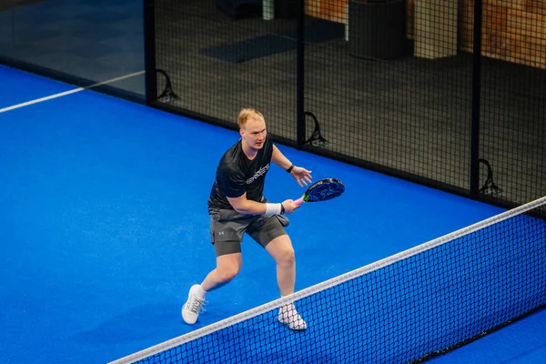 Riga, Letonya - 24 Ocak 2026: Padel turnuvası. Gri şortlu ve siyah tişörtlü erkek padel oyuncusu mavi sahada net ve duvarlarla topa vuruyor.