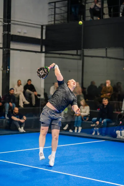 Riga, Letonya - 24 Ocak 2026: Padel turnuvası. Siyah tişörtlü ve şortlu bir erkek atlet, mavi bir padel kortunda seyircilerin izlediği bir topa servis yapıyor.