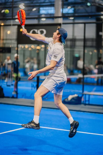 Riga, Letonya - 24 Ocak 2026: Padel turnuvası. Gri elbiseli erkek sporcu, canlı bir mavi sahada topa vurmak için atlıyor.