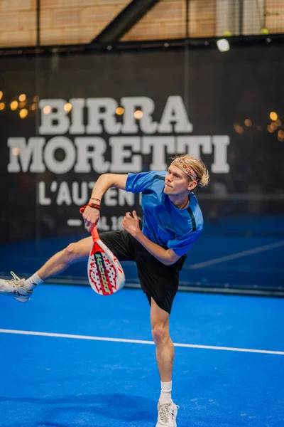 Riga, Letonya - 24 Ocak 2026: Padel turnuvası. Mavi tişörtlü ve siyah şortlu genç padel oyuncusu mavi kortta topa vurmaya hazırlanıyor.