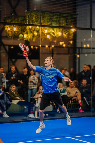 Riga, Letonya - 24 Ocak 2026: Padel turnuvası. Mavi tişörtlü ve siyah şortlu erkek padel oyuncu kapalı sahada seyirciler izlerken topa vurmak için zıplıyor.