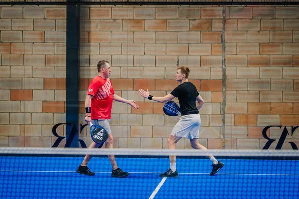 Riga, Letonya - 24 Ocak 2026: Padel turnuvası. Spor kıyafetli iki adam, modern bir bina içinde mavi bir padel kortunda birbirlerini selamlıyorlar.