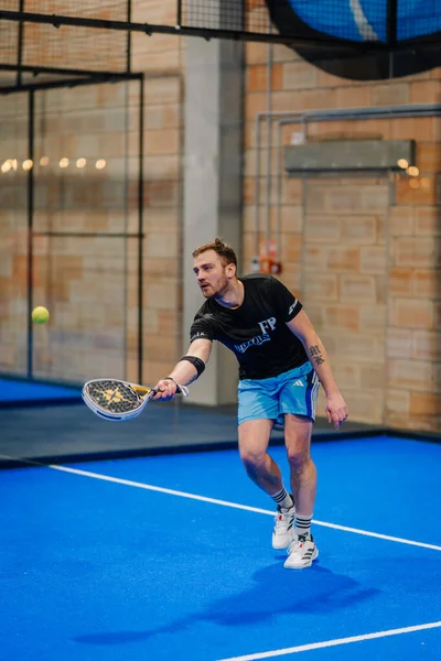 Riga, Letonya - 24 Ocak 2026: Padel turnuvası. Siyah ve mavi giysili erkek padel atlet canlı mavi bir iç sahada topa vurmaya hazırlanıyor.