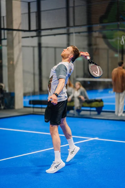 Riga, Letonya - 24 Ocak 2026: Padel turnuvası. Spor giyim sektöründe erkek atlet, arka planda oyuncular ve izleyicilerle mavi sahada bir padel topu sunuyor.