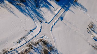 Kesişen yollar ve ağaç gölgelerinin yer aldığı karlı bir arazinin hava perspektifi, kış manzarasının sükunetini vurguluyor