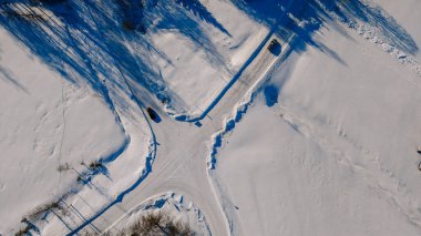Karlı bir arazinin hava perspektifi kışın etrafını saran ağaçların gölgeleri ve görünen lastik izleriyle yol ayrımını gösteriyor.