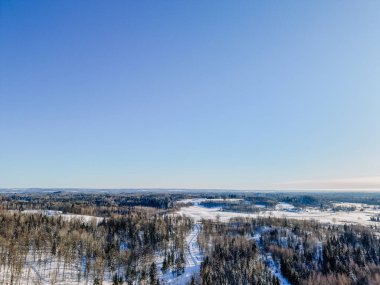 Karla kaplı zemin, yemyeşil ağaçlar ve açık mavi gökyüzünün altında dolambaçlı yollar olan kış manzarası, doğal güzellikleri vurguluyor.