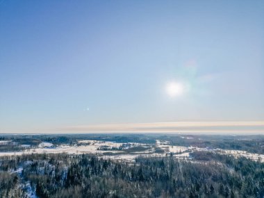 Açık mavi gökyüzünün altında, karla kaplı tarlalar ve her zaman yeşil olan ormanlar manzarayı aydınlatan parlak bir güneşle dolu bir kış manzarasının havadan bakış açısı.