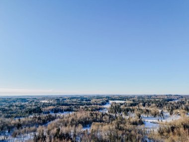 Parlak mavi gökyüzü altında karla kaplı tarlalar ve sık ormanların yer aldığı kış manzarasına ait hava perspektifi doğanın güzelliğini gözler önüne seriyor.