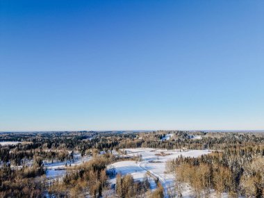 Parlak mavi gökyüzü altında karla kaplı tepeler ve yemyeşil ormanların yer aldığı bir kış manzarasına ait hava perspektifi, dingin ortamı aydınlatıyor.
