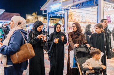 Dubai, Birleşik Arap Emirlikleri - 13 Ocak 2026: Siyah abayalar içindeki kadınlar Dubai Global Village 'da abur cubur gezintisi yapıyorlar, arka planda görünen dükkanlar aydınlatılıyor