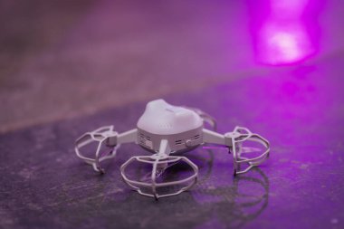 Riga, Letonya - 30 Ocak 2026: Şeker Diyarı. Beyaz kuadkopter İHA 'sı mor çevre aydınlatmasının altında parlak bir yüzeye yerleştirilmiş güvenlik kafesi ile