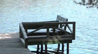 Sakin suyun üzerindeki bir rıhtımda ahşap bir bank, arka planda yansımalar ve ağaçlar, rahatlamak için sakin bir açık hava ortamı yaratıyor.