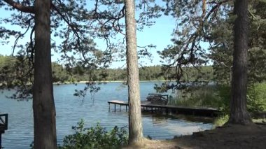 Uzun ağaçlar ve yeşilliklerle çerçevelenmiş sakin göl manzarası. Parlak mavi gökyüzünün altında durgun suya uzanan ahşap bir rıhtım var.
