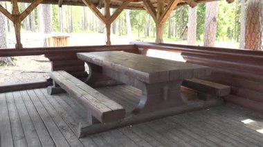Ağaçlar ve doğal elementlerle çevrili bir ormanda, kırsal bir taraçanın altında bulunan ahşap piknik masası ve banklar, sakin bir açık hava alanı yaratıyor.