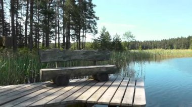 Sakin bir gölün yanındaki rıhtımda bulunan ahşap bir bank. Arka planda ağaçlar ve çimenler var. Huzurlu ve huzurlu bir doğal atmosfer yaratıyor.
