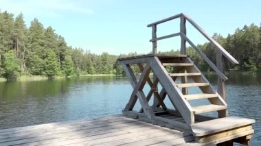 Tahta adımlar, yeşil ağaçlar ve parlak gökyüzü tarafından çerçevelenmiş sakin bir göle yükselir ve doğada huzurlu bir geri çekilme sağlar.
