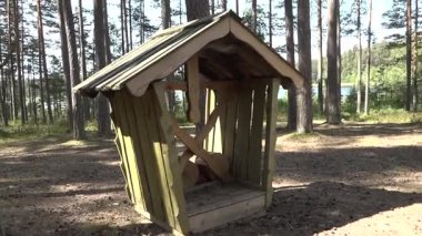 Ahşap bir sığınak eğimli bir çatı, bir orman açıklığı, uzun ağaçlar ve arka planda sakin bir göl ile çevrili