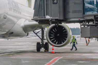 Riga, Letonya - 06 Şubat 2026: Güvenlik ekipmanlı uçak teknisyeni, yolcu köprüsü görünen havaalanı terminalindeki jet motorunu inceliyor