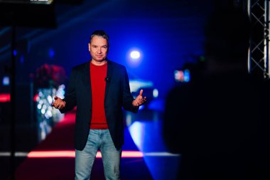 Riga, Letonya - 4 Şubat 2026: Chery. Sahnede siyah ceketli ve kırmızı gömlekli bir sunucu parlak ışıklarla ve arka planda bir araçla sahneye çıkıyor.