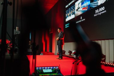 Riga, Letonya - 4 Şubat 2026: Chery. Sahnede Chery TIGGO modellerini sunan konuşmacı, marka ayrıntılarını ve izleyici üyelerini gösteren geniş bir ekran sunuyor.