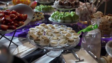 Tatlı masasında çeşitli tatlılar sergilenir, kapkekler, badem kurabiyeleri ve çilekler, mor bir masa örtüsü ile güzel bir şekilde cam tabaklar üzerinde sıralanmış.