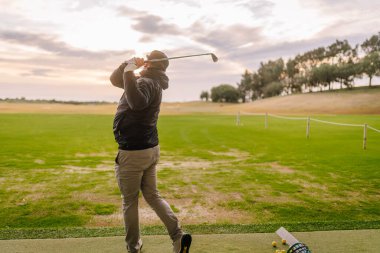 Siyah ceketli bir adam golf sopasıyla golf sopasını alıp yeşil çimlerle ve bulutlu bir gökyüzünün altındaki ağaçlarla çevrili bir alanda sallıyor.