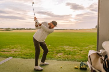 Erkek golfçü golf sahasında golf topuna vurmaya hazırlanıyor. Arka planda çimenli bir alan ve bulutlu bir gökyüzü var.