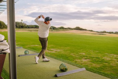 Uzun kollu beyaz gömlekli ve şapkalı bir adam golf sahasındaki golf topuna vuruyor, arka planda yemyeşil çimenler ve ağaçlar var.