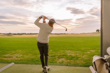 Erkek golfçü golf sopasıyla golf sahasında, yeşil çimlerle çevrili ve arka planda bulutlu bir gökyüzü ile atış yapar.