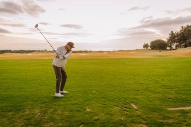 Beyaz kazaklı ve şapkalı bir adam yemyeşil bir sahada golf sopası sallamaya hazırlanıyor. Gün batımında ağaçlar ve tepeler görülebiliyor.