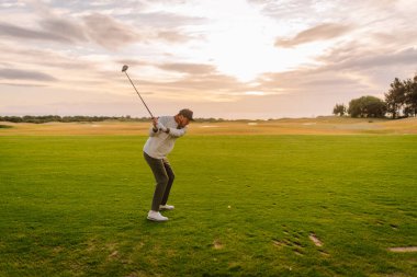 Erkek golfçü, günbatımında yemyeşil bir sahada geniş bir alan ve arka planda ağaçlarla golf topuna vurmaya hazırlanıyor.