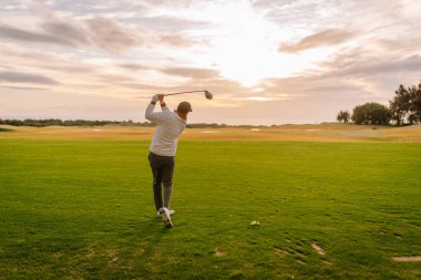 Beyaz, uzun kollu gömlekli bir adam gün batımında yemyeşil bir çayırda golf salıncağına biniyor. Ağaçları ve arka planda açık manzarası var.