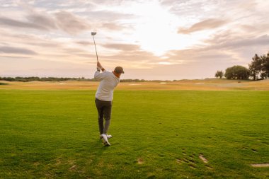 Erkek golfçü, arka planda altın çimenler ve ağaçlarla çevrili, yemyeşil bir sahada golf topuna vurmaya hazırlanıyor.