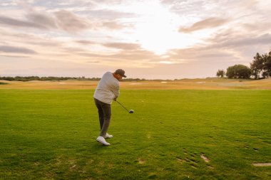Erkek golfçü, günbatımında yemyeşil bir sahada, arka planda ağaçlar ve açık alanlar olan bir golf topuna vurmaya hazırlanıyor.