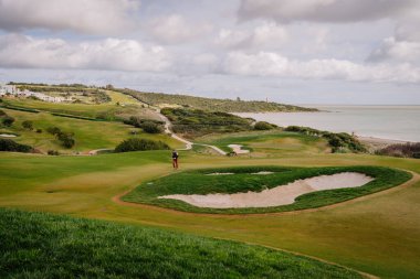 Golf course scene with a golfer standing near a sand trap, surrounded by lush green hills and an ocean view under a cloudy sky