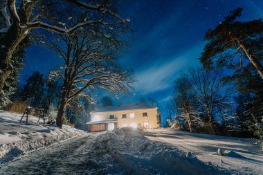 Geceleri karlı bir arazide aydınlık bir ev, dolambaçlı bir garaj yolu ve yıldızlı bir gökyüzünün altında uzun ağaçlar, sakin bir kış manzarası yaratıyor.